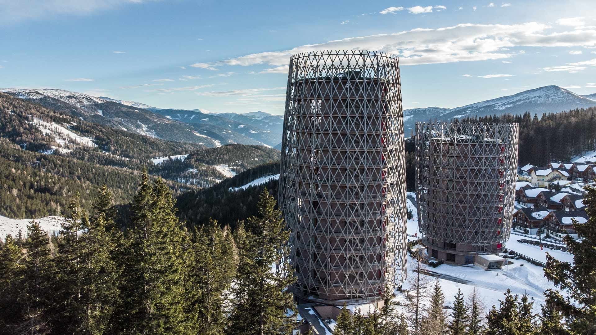 Two cylindrical buildings with lattice-like exteriors stand in a snowy mountain landscape with evergreen trees.