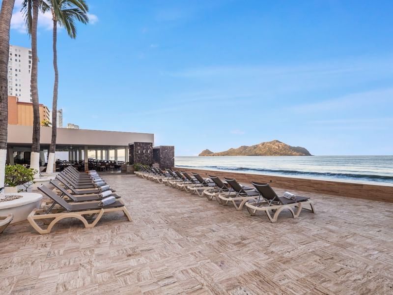 Oceanfront patio with rows of lounge chairs overlooking the sea & a distant island under a clear sky at Sunvivia Mazatlán