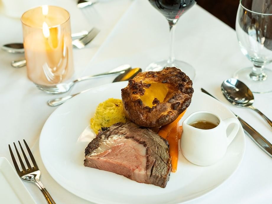 Plate with beef, potatoes, vegetables, gravy, and Yorkshire pudding, surrounded by cutlery and glasses on a table.