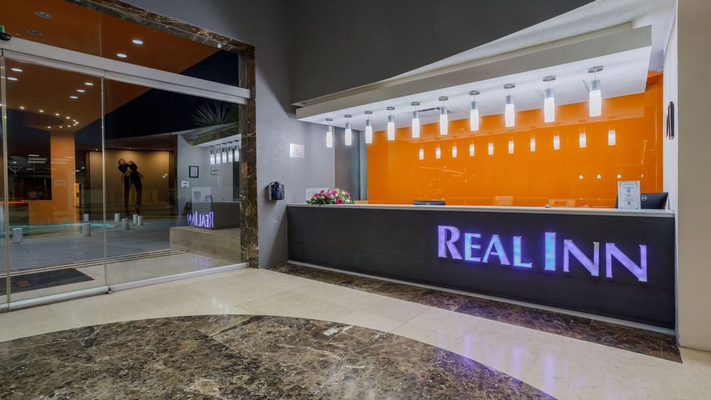 Night view of the reception showing the glowing logo and vibrant orange glass wall at Real Inn Torreon