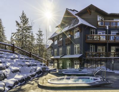 Blackstone Mountain Lodge surrounded by snow-covered trees, with a steaming hot tub under the bright sun