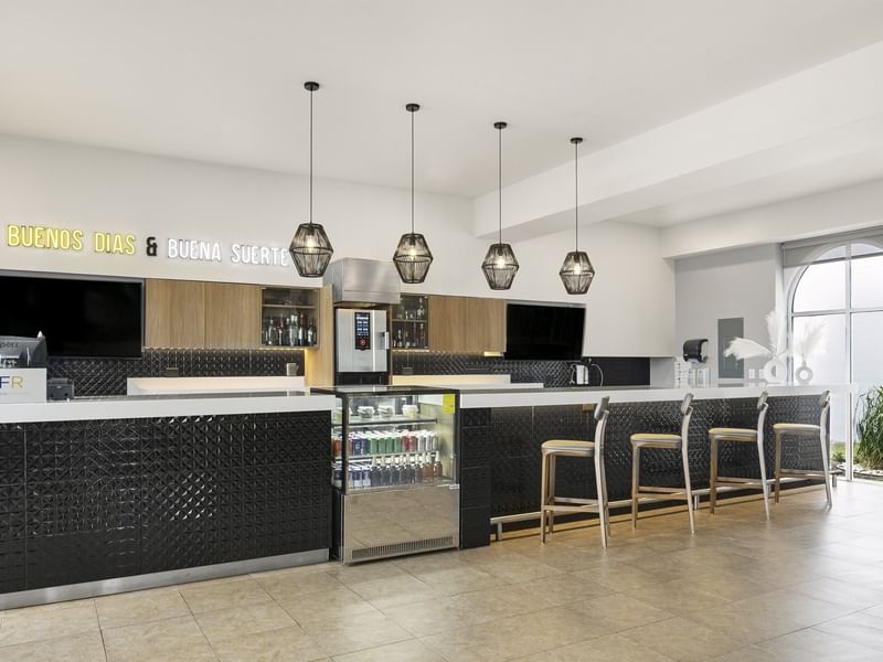 Stylish lobby bar at Fiesta Inn Express Nuevo Laredo with pendant lights, a cooler, and a neon sign reading Buenos Días