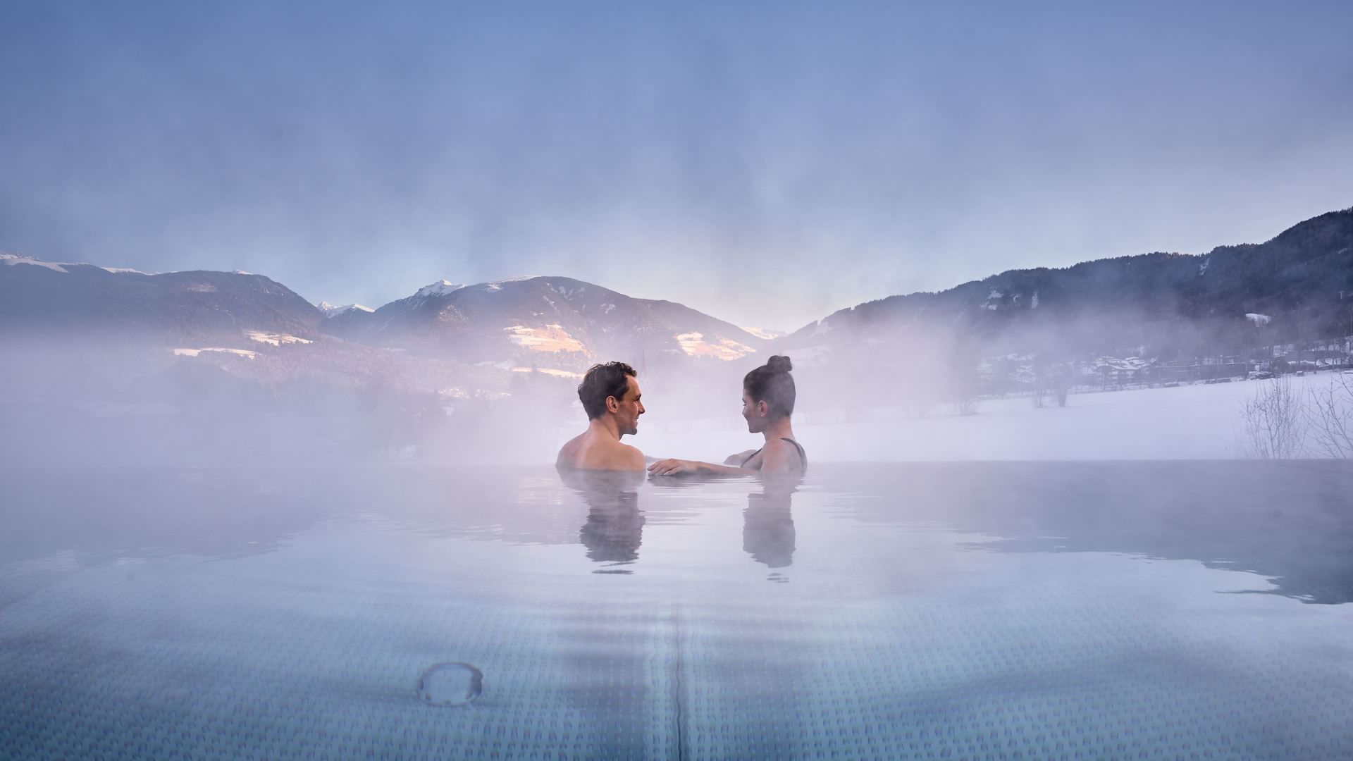 Couple relaxing in a misty pool by the mountains at Falkensteiner Hotel Kronplatz