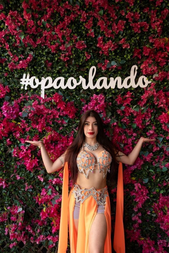 A woman in an orange belly dancing costume stands in front of a pink floral wall with a sign reading OPA ORLANDO.