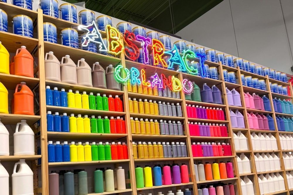 A large wall with containers of colorful paint and a glowing neon sign that says Abstract Orlando.