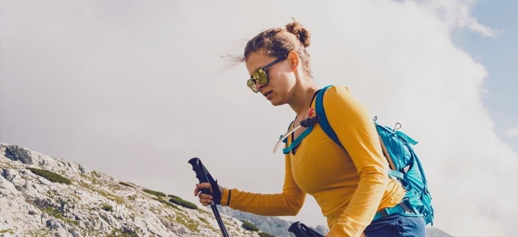 A woman in a bright yellow long-sleeve shirt and sunglasses hikes uphill with trekking poles, carrying a blue backpack, against a backdrop of rocky slopes and cloudy sky.