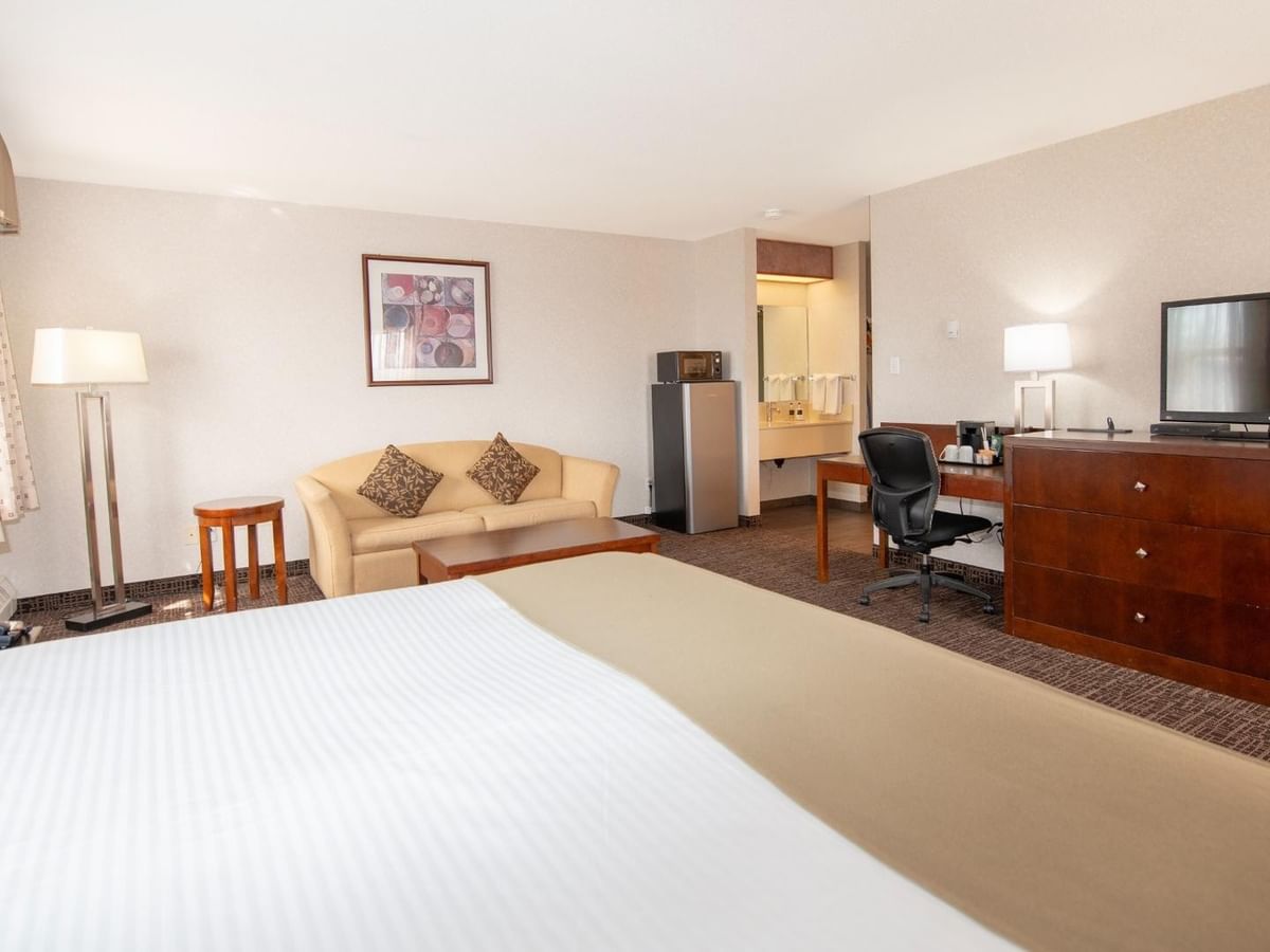 A hotel room with a bed, couch, TV, desk, refrigerator, and artwork on the wall.