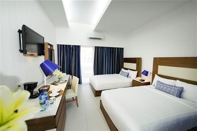 A hotel room at Hotel Mykonos with two full-sized beds, blue curtains and a modern desk area