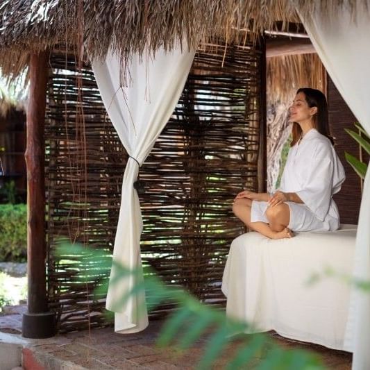 Mujer en traje de spa, meditando en cama bajo dosel en cabaña al aire libre.