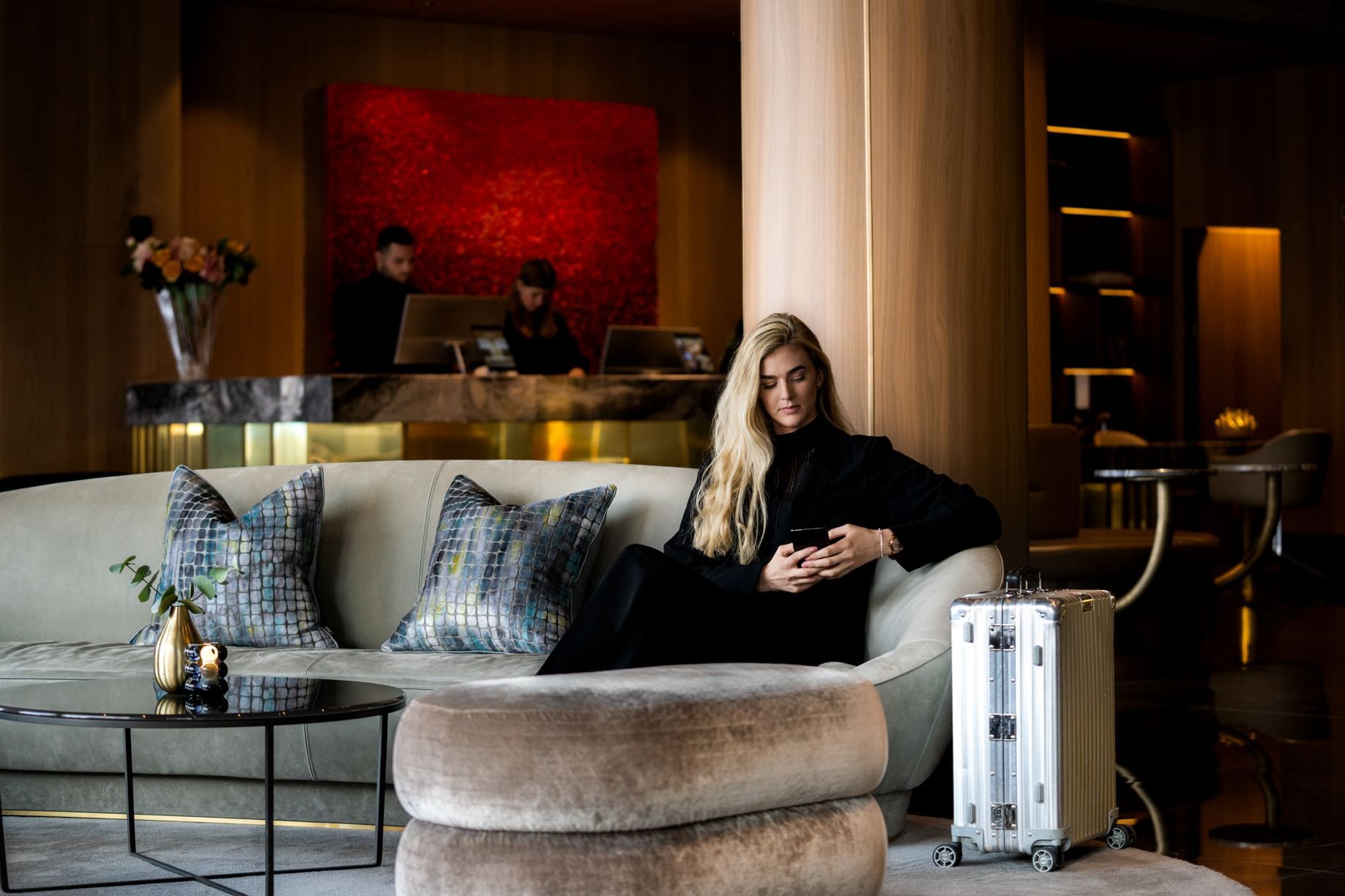 Blond kvinna sitter på soffa med kuddar och väska, två personer bakom räknare med datorer.