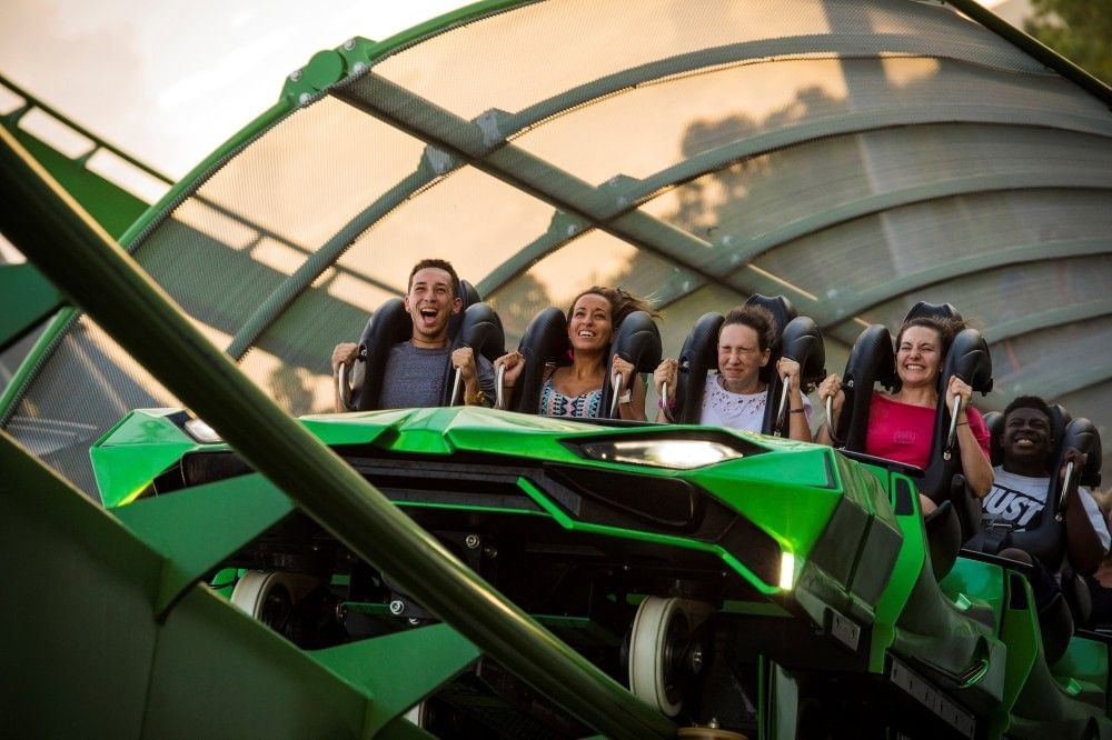 A row of adults screaming and smiling on a green roller coaster car, about to launch. 