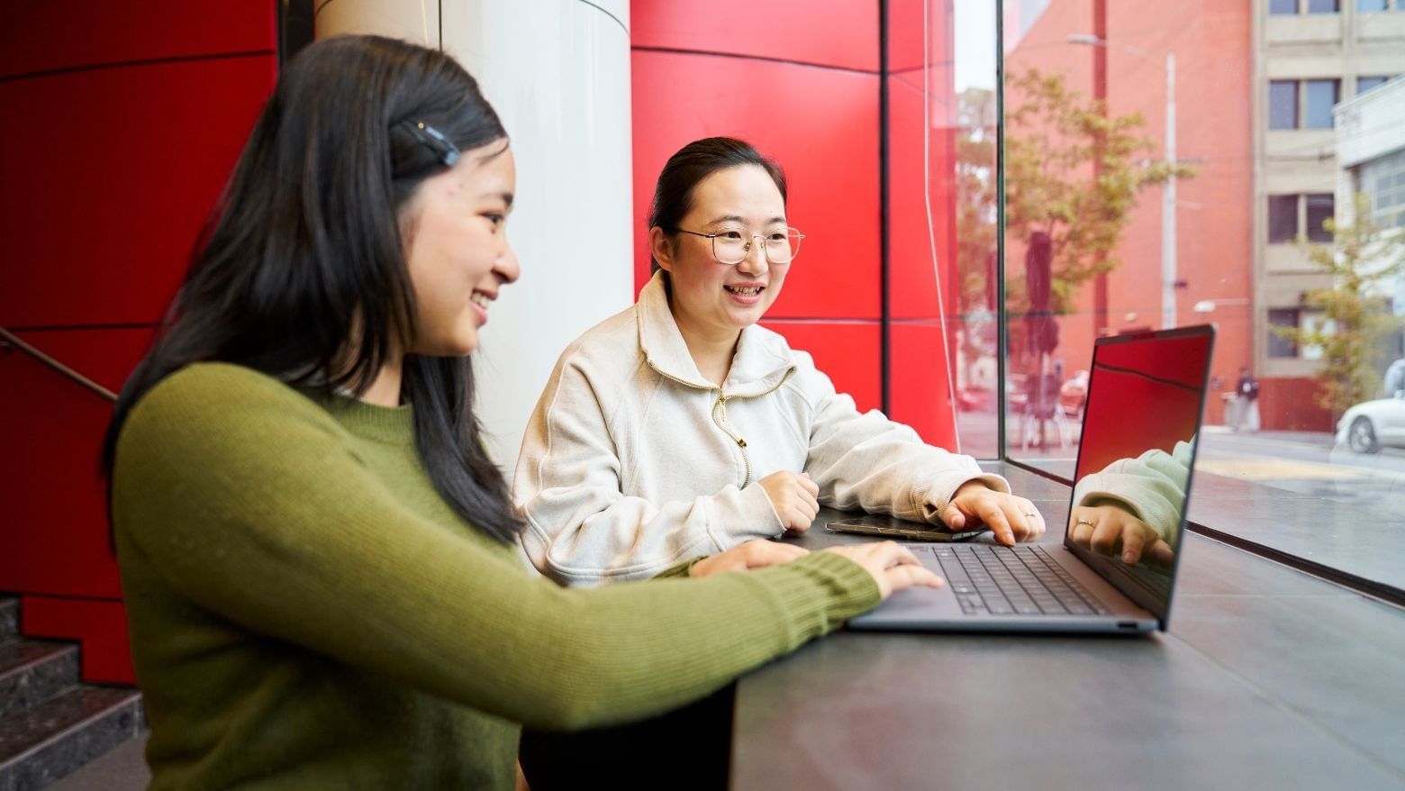 Two residents working on a laptop with a streetscape view in the background at Student Living D2.