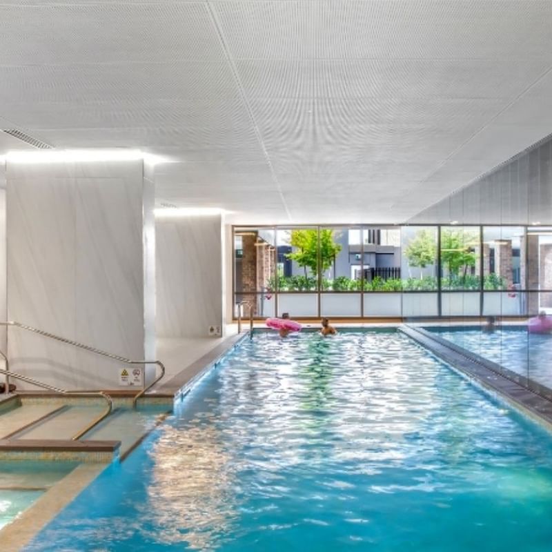 Indoor pool area with blue water, glass walls, and people relaxing in the pool.