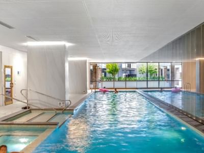 Indoor pool area with blue water, glass walls, and people relaxing in the pool.