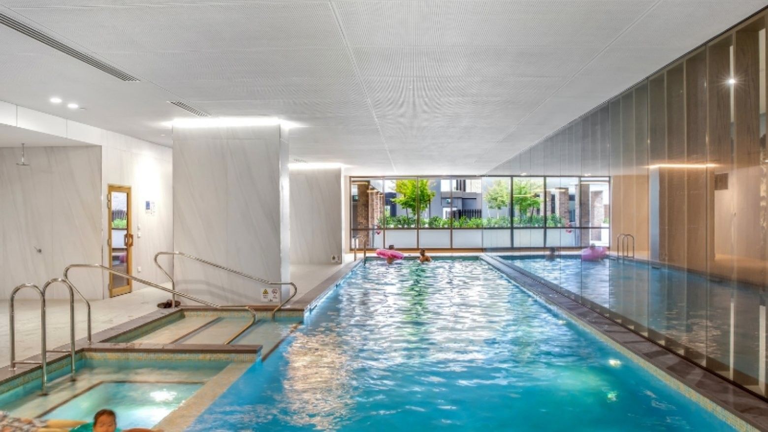 Indoor pool area with blue water, glass walls, and people relaxing in the pool.