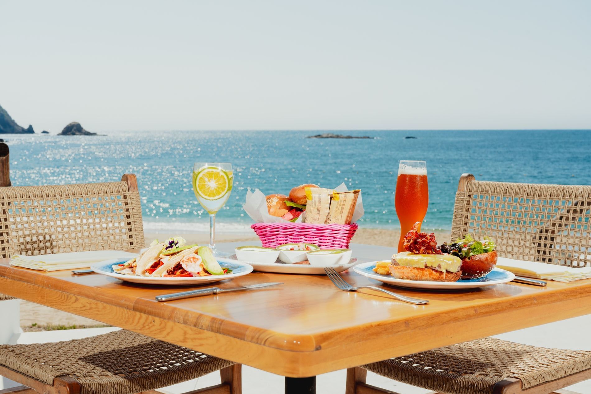Wooden dining table with four wicker chairs on a sunny beach at Bel La Grill in Camino Real Zaashila Huatulco