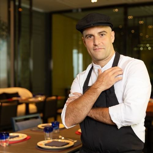 A chef in a black apron posing in front of a restaurant.