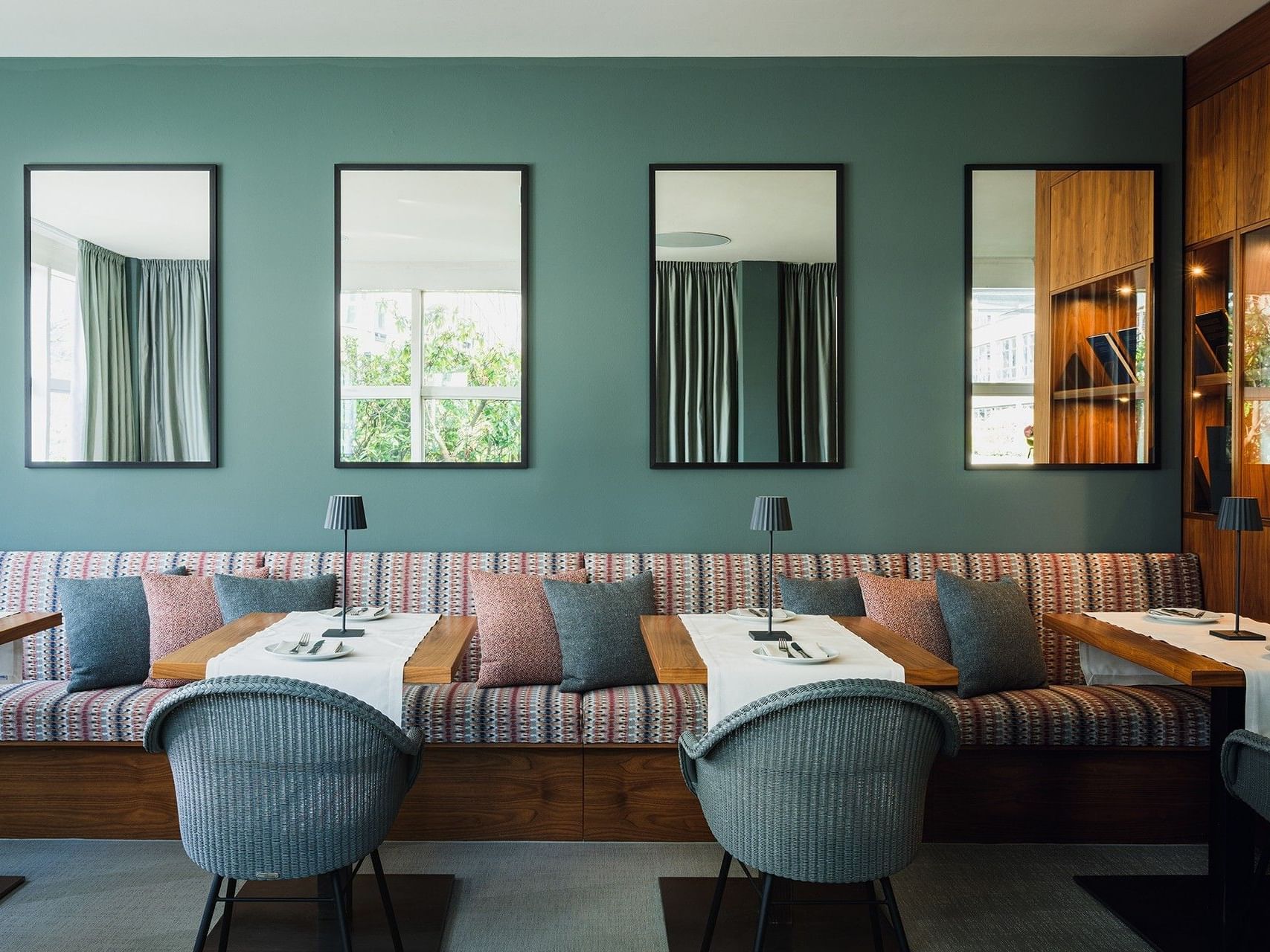 Grün gestrichene Wand mit vier Spiegeln, einer langen Bank mit Kissen und drei Tischen mit Stühlen und Lampen.