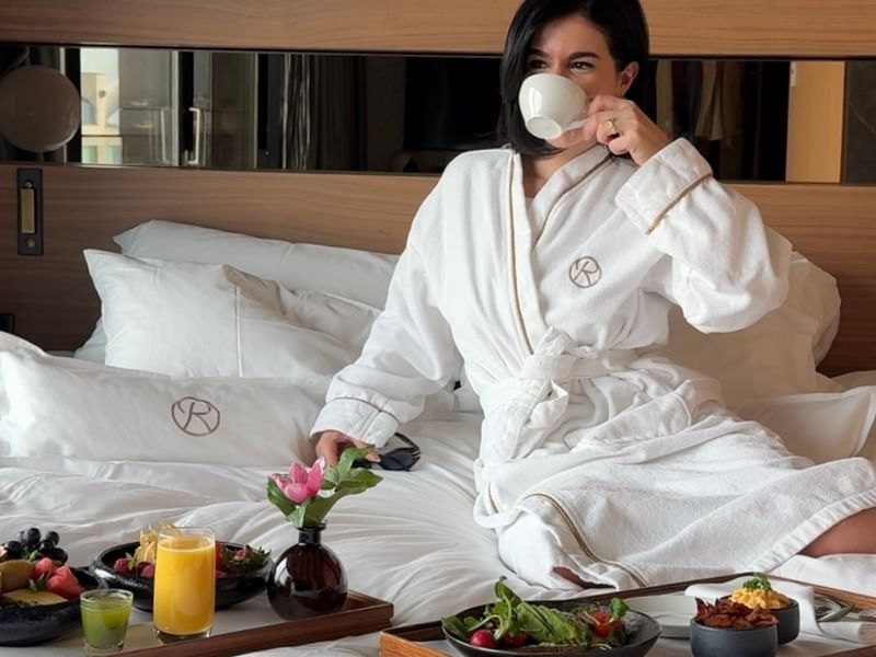 Kvinde i badekåbe drikker kaffe i sengen med morgenmad i Stay Four Longer-tilbuddet.