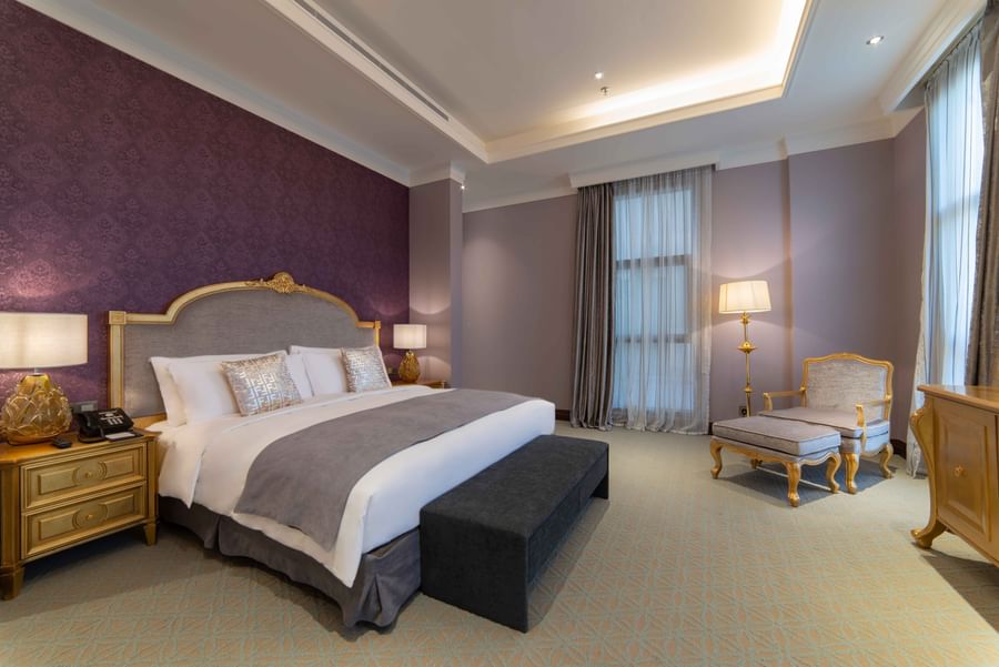 Plush bed with a gold headboard by a chair and footrest under purple walls in Penthouse Villa at Warwick Al Khobar