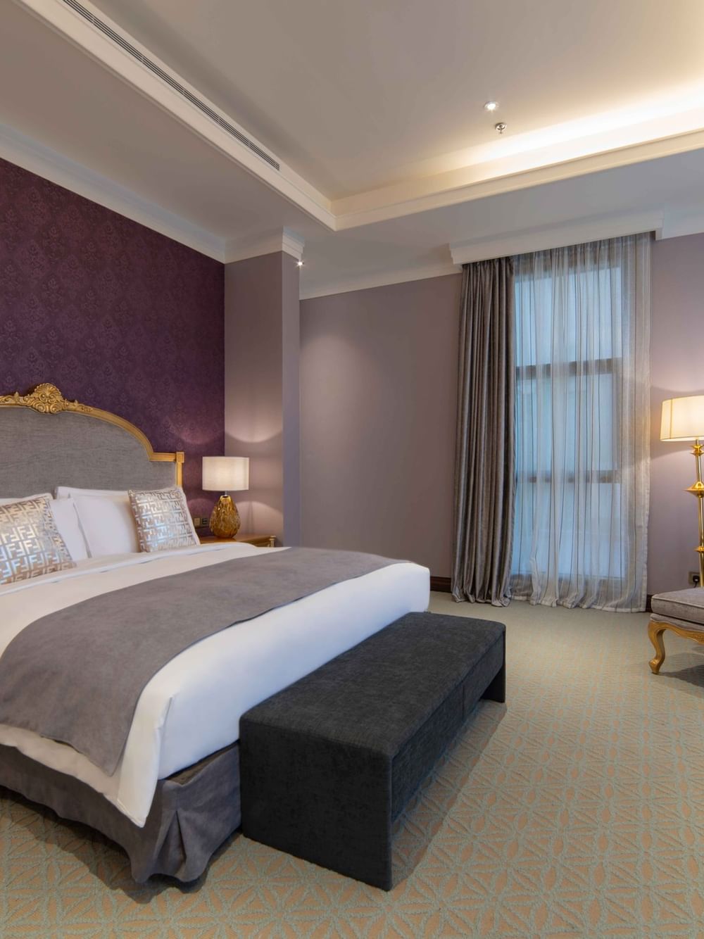 Plush bed with a gold headboard by a chair and footrest under purple walls in Penthouse Villa at Warwick Al Khobar