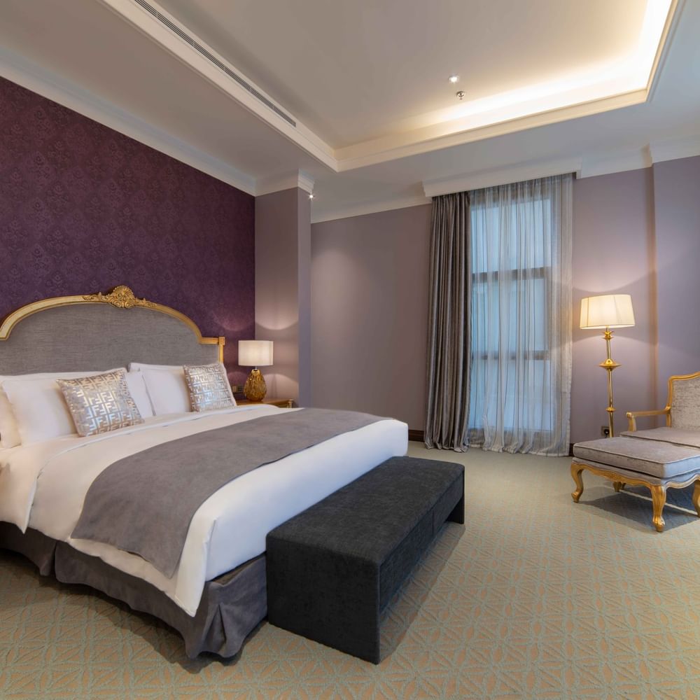 Plush bed with a gold headboard by a chair and footrest under purple walls in Penthouse Villa at Warwick Al Khobar