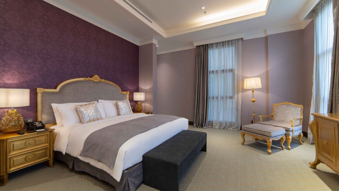 Plush bed with a gold headboard by a chair and footrest under purple walls in Penthouse Villa at Warwick Al Khobar