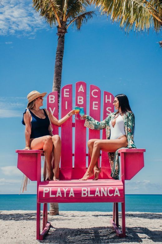 Ladies posing on a big Beach Vibes chair with cocktails at Playa Blanca Beach Resort