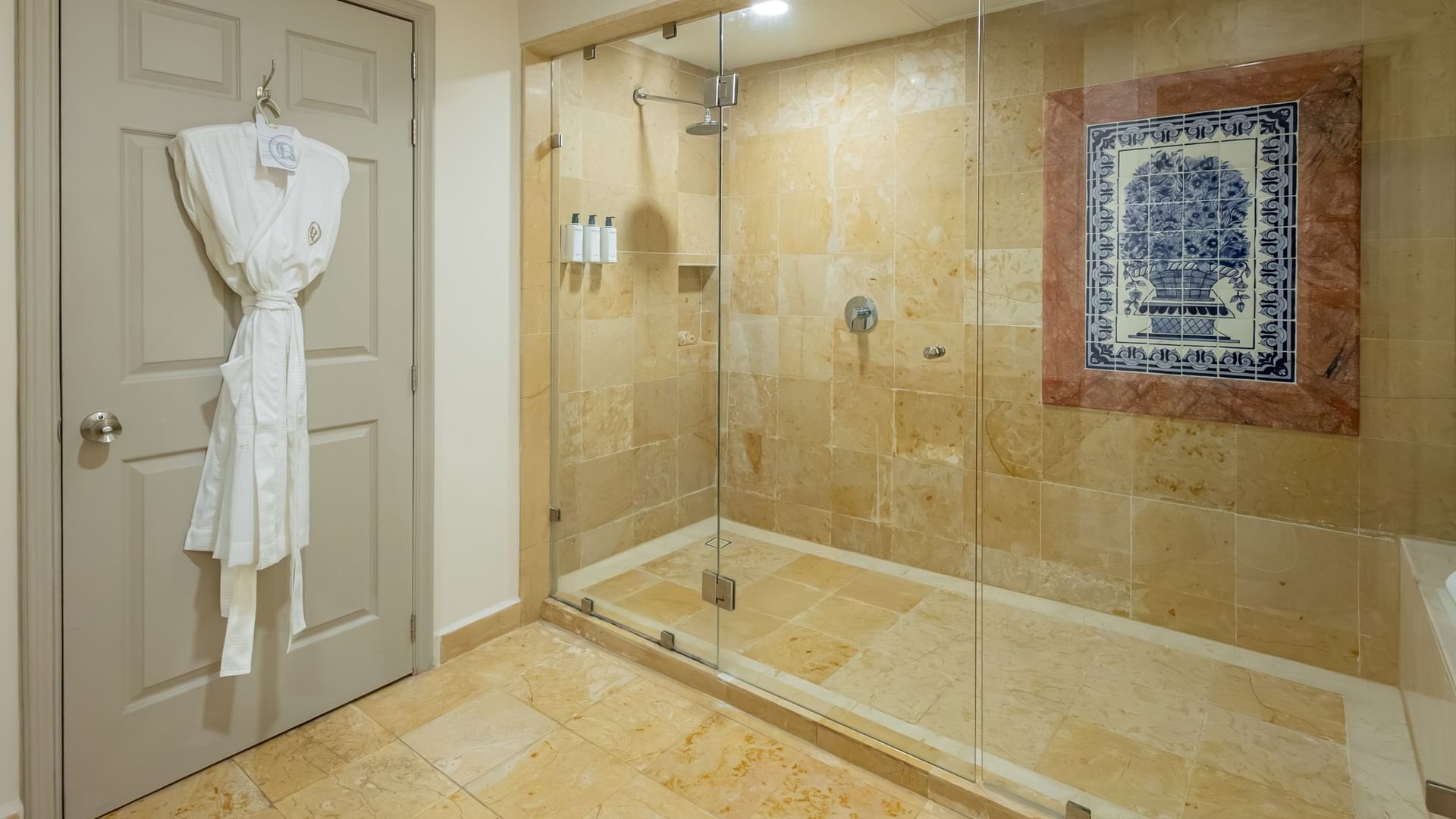 Baño con ducha de cristal, azulejo beige, bata blanca y decoración de talavera azul en Quinta Real Monterrey.