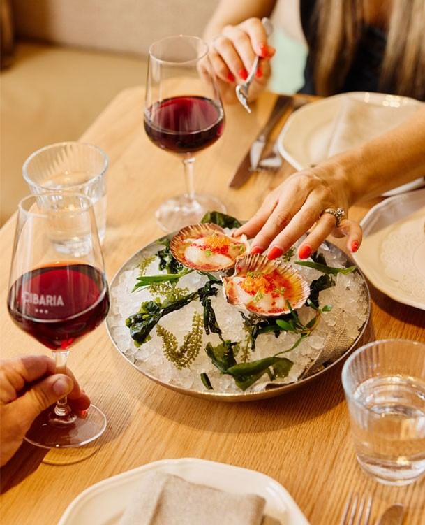 A woman tasting seared scallops on ice with a glass of red wine at Elysium Hotels