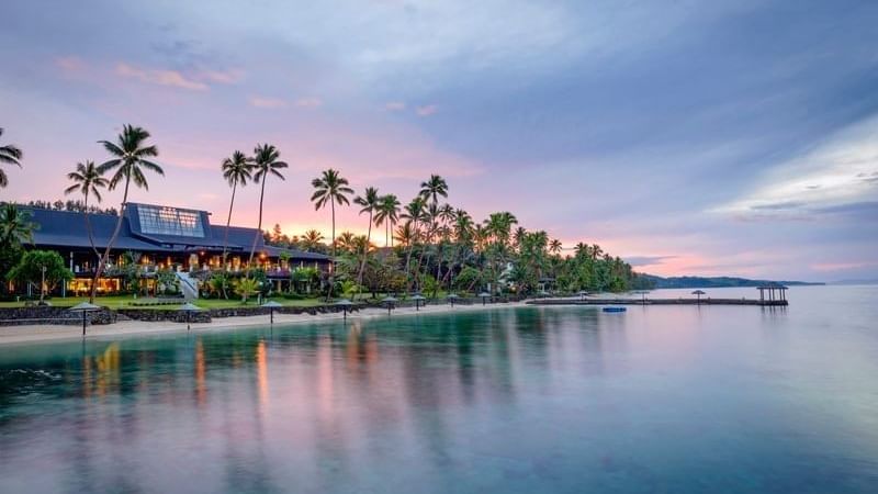 Sunset over a tranquil ocean at Warwick Fiji resort, with palm trees lining the sandy beach