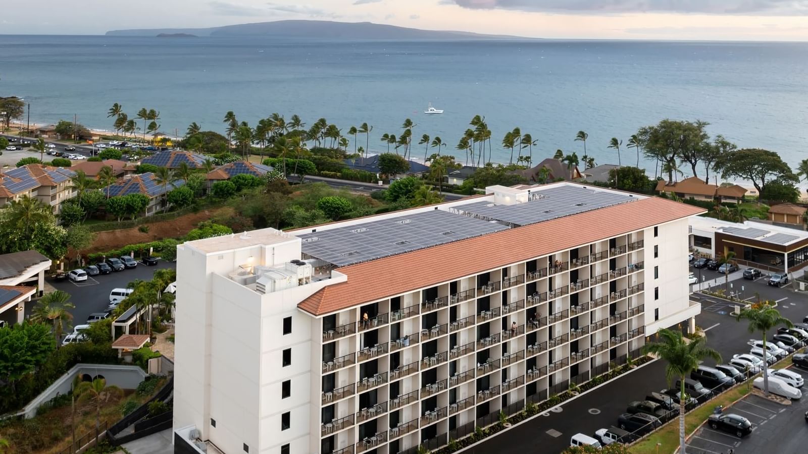 Aerial view of a white, multi-story building with balconies and solar panels on the roof, surrounded by a parking lot and tropical landscape.