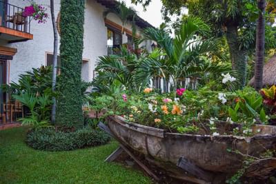 Wooden boat planter filled with colorful flowers in lush garden beside a white building with balconies.