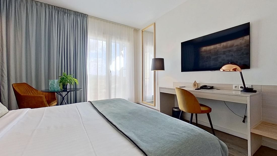 Modern hotel room with a bed, desk, chair, TV, and window with curtains.