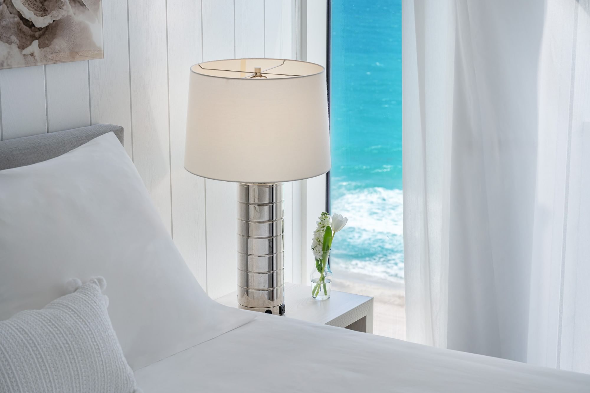 A close-up of a bed with white bedding and a lamp on a nightstand, with a view of the ocean through a window.