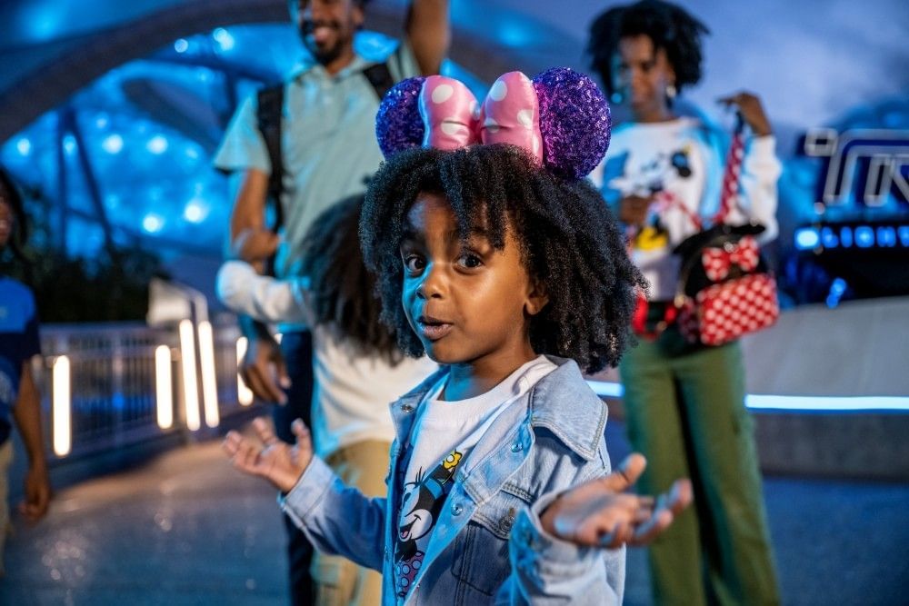 A toddler girl in a denim jacket shrugs at the camera, her parents standing behind her in front of the Tron ride at Magic Kingdom.