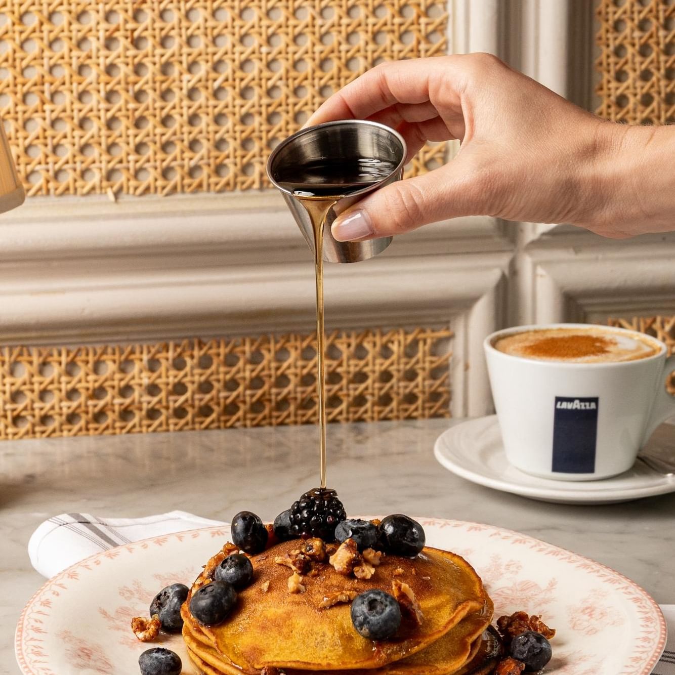 Hand pouring syrup over golden pumpkin pancakes topped with fresh berries and walnuts beside a coffee cup.