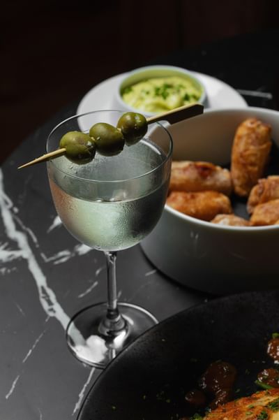 Cocktail glass with three olives on a skewer, served alongside golden fried snacks and green dipping sauce at Cypress & Oak