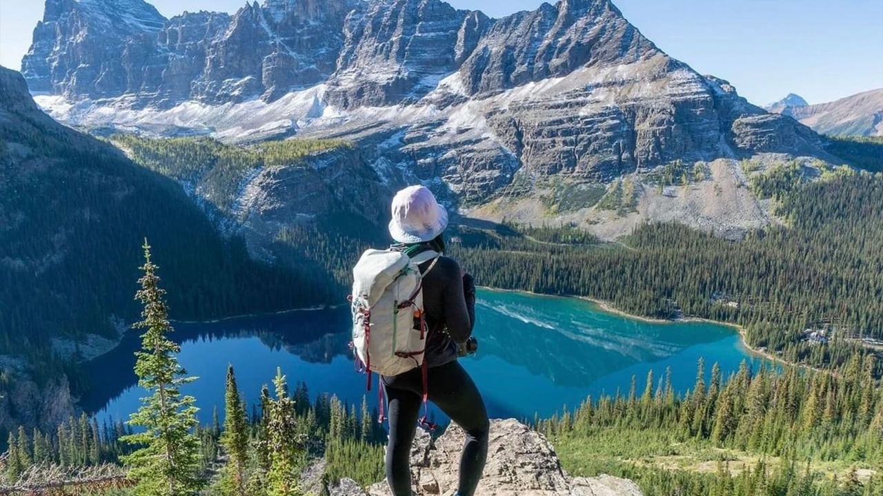 A hiker with a backpack stands on a rock overlooking a mountain lake and lush forests.