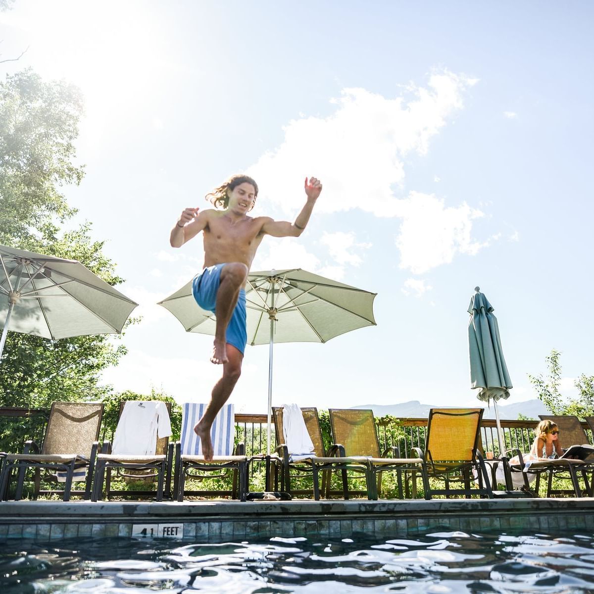 Young boy jumping into an outdoor pool surrounded by lounge chairs and umbrellas on a sunny summer day.