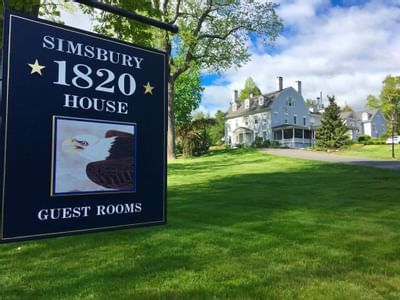 Exterior view of Simsbury 1820 House with a guest room sign under a tree, by a lawn and gray colonial building beyond