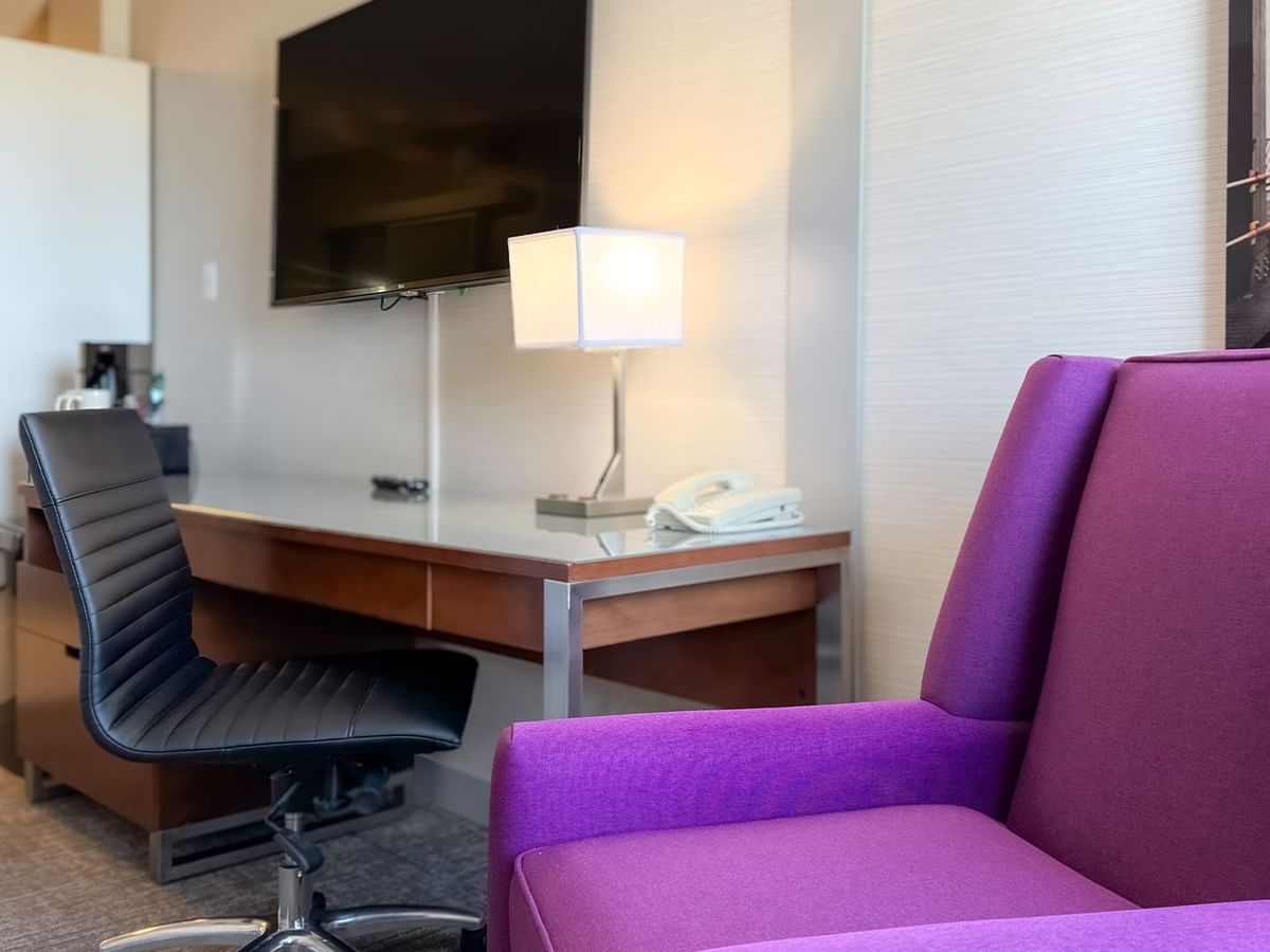 Hotel room with work desk, purple armchair, lamp, TV, telephone, and artwork.