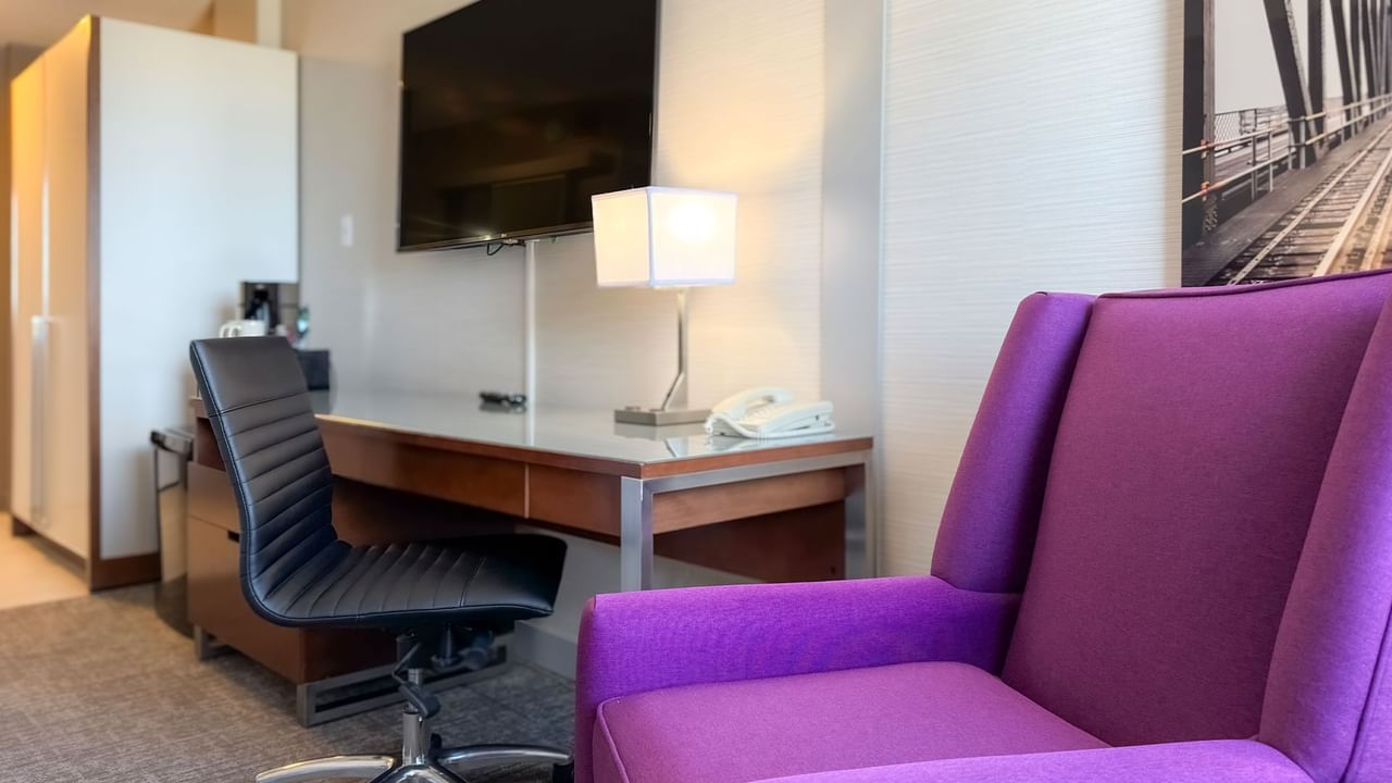 Hotel room with work desk, purple armchair, lamp, TV, telephone, and artwork.