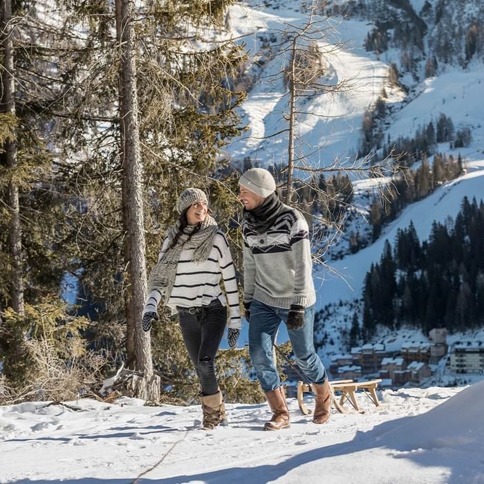 Zwei Menschen spazieren auf einem schneebedeckten Pfad in den Bergen, umgeben von Bäumen.