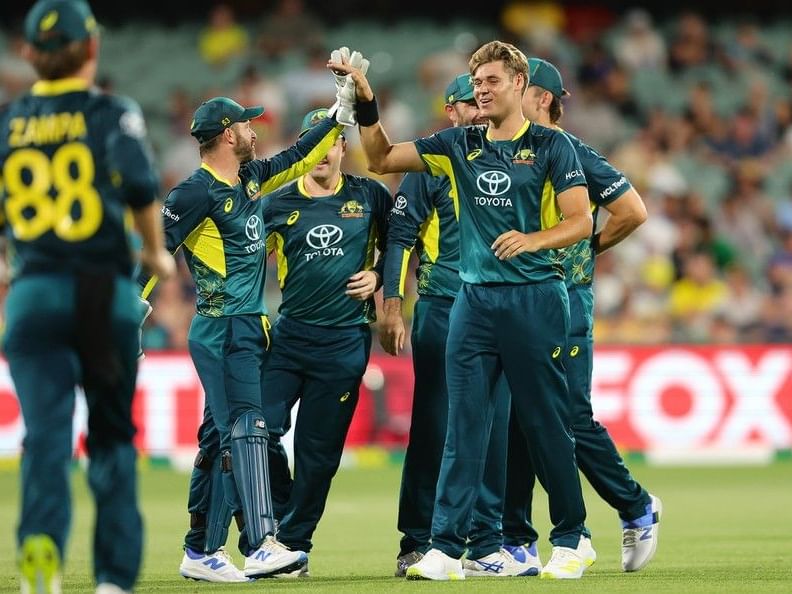 Cricket players in green and yellow uniforms celebrate on a field with a crowd in the background.