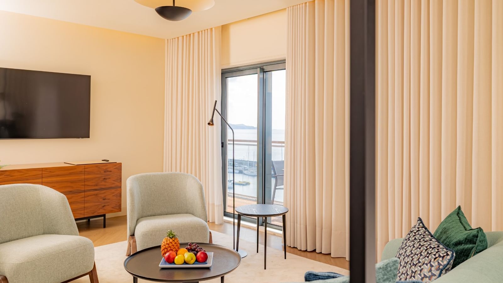 Sala de estar com sofá, cadeiras, mesa de café com frutas e TV na parede.