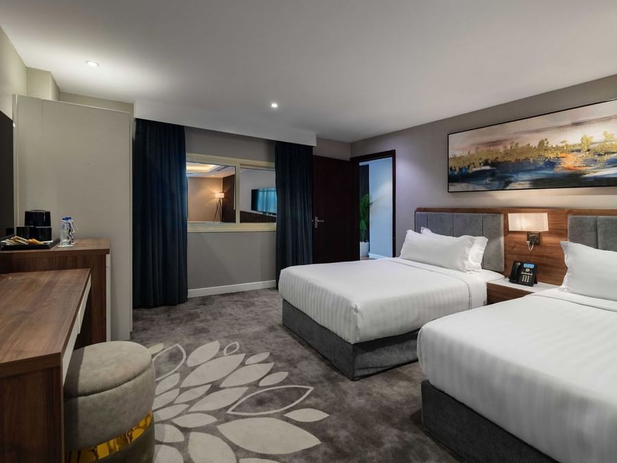 Two white striped beds beside a wooden desk and a round stool under a wall mirror in Standard Room at Warwick Al Jubail