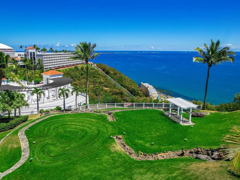 Aerial view with a lush lawn, white pergola, palm trees, and Las Casitas Village overlooking the blue ocean