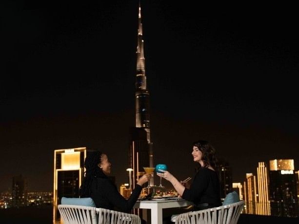 Two women toasting with cocktails at a rooftop bar at Paramount Hotel Midtown, with the Burj Khalifa in the background