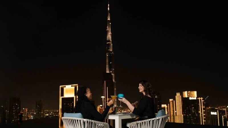 Two women toasting with cocktails at a rooftop bar at Paramount Hotel Midtown, with the Burj Khalifa in the background